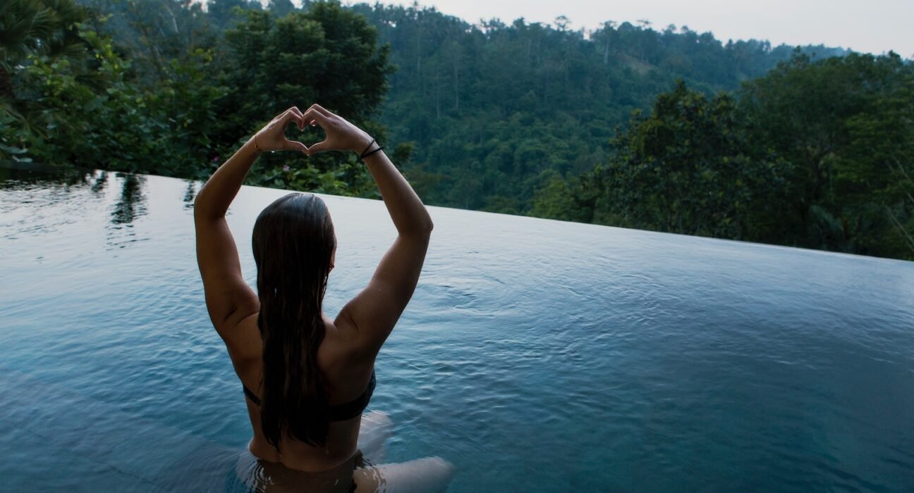 woman in infinity pool making heart hand gesture facing green leafed trees