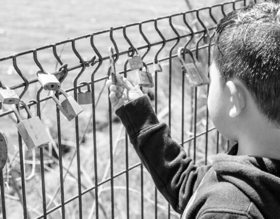 grayscale photography of boy holding padlock on fence